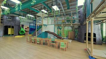 A spacious indoor play area with a wooden floor, a slide, climbing structures, and a seating area with chairs and tables.