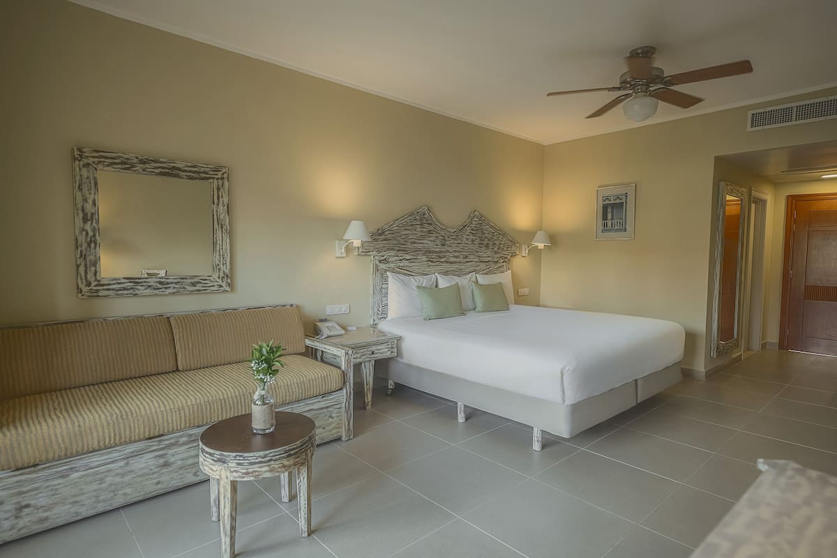Une chambre d’hôtel avec un grand lit, un canapé, une petite table avec une plante, un ventilateur de plafond et un miroir au mur.