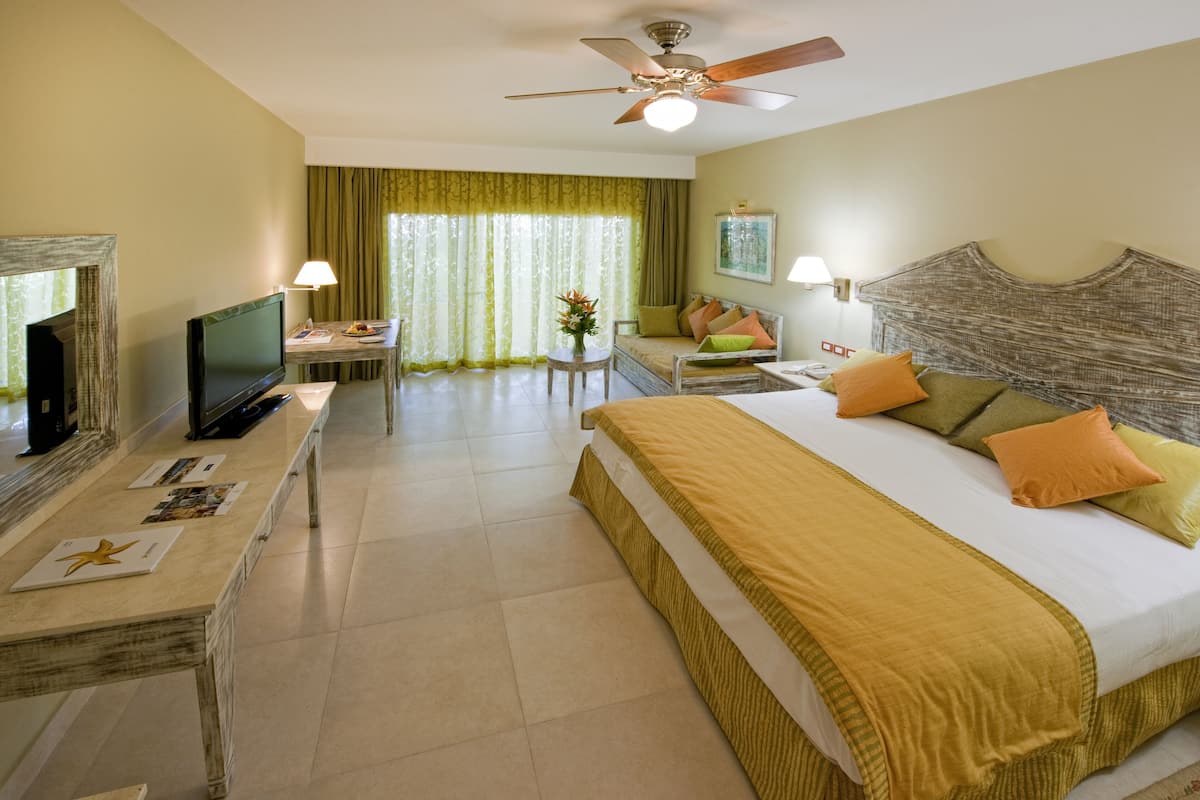 Une chambre d’hôtel avec un lit, un bureau avec une télévision, un ventilateur de plafond et une fenêtre avec des rideaux.