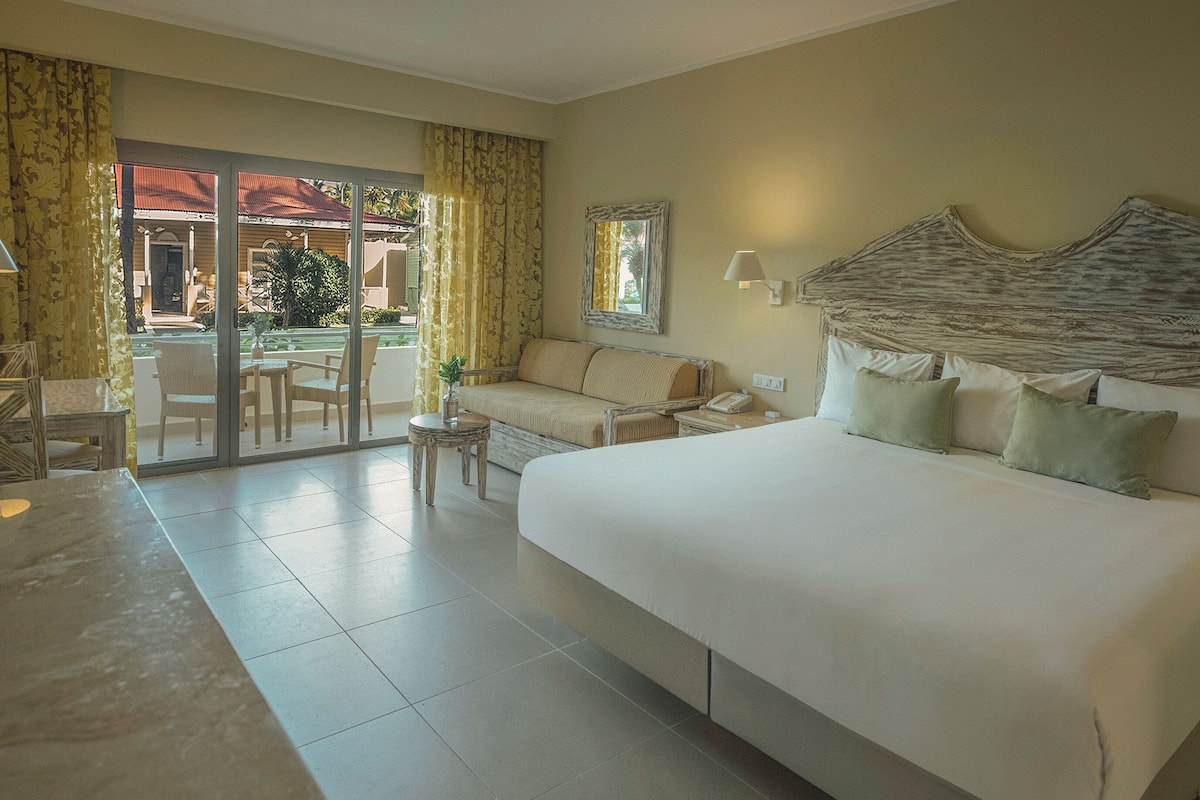 Une chambre d’hôtel avec un grand lit, un canapé, une petite table et une chaise. Une porte-fenêtre coulissante donne sur un espace extérieur agrémenté d’une piscine et de palmiers.