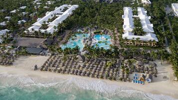An aerial view of a resort with multiple buildings, a central swimming pool, and a beach area with umbrellas and lounge chairs.