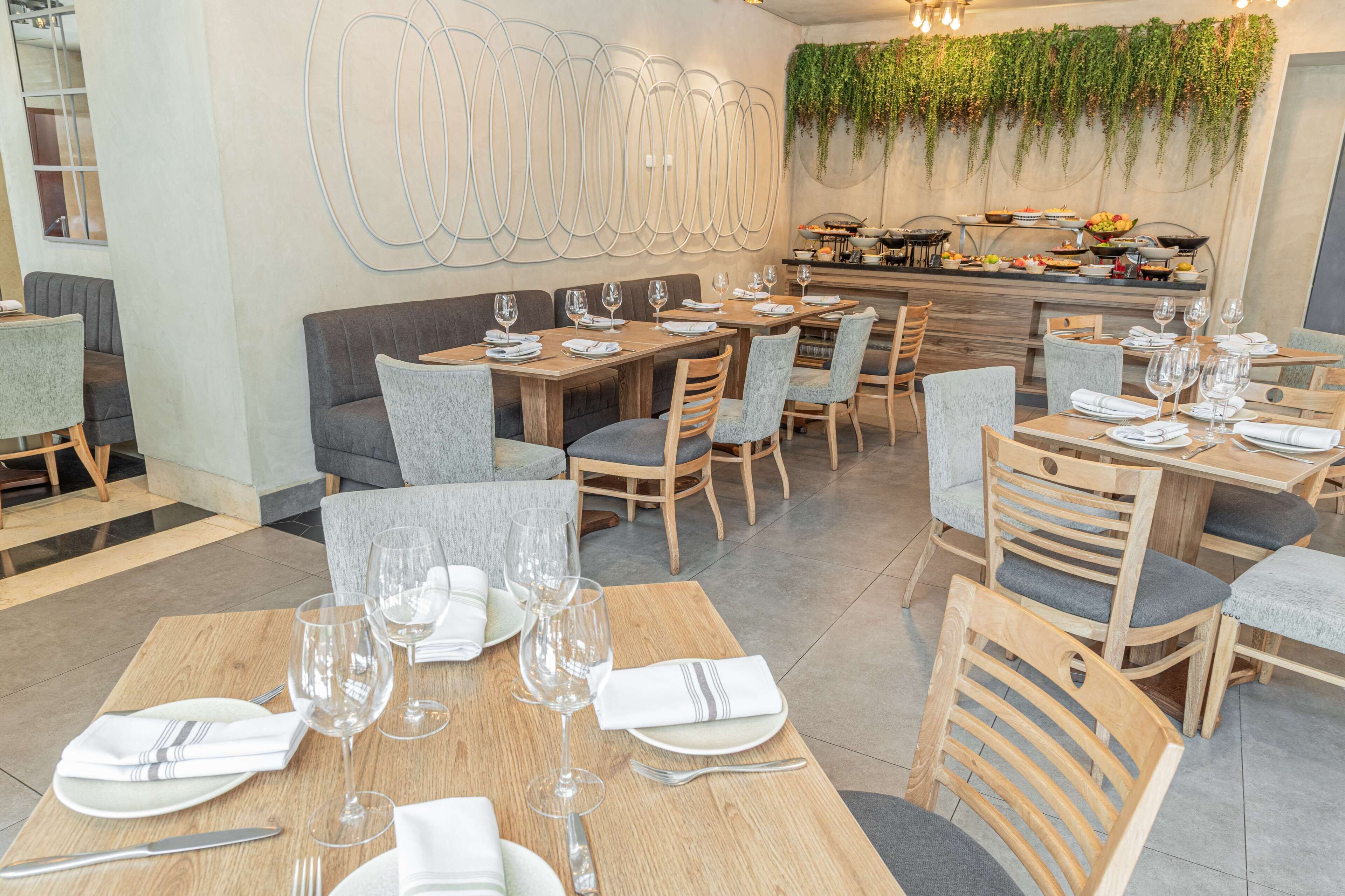 A restaurant with wooden tables and grey upholstered chairs, a wall-mounted abstract art piece, and a green plant wall.