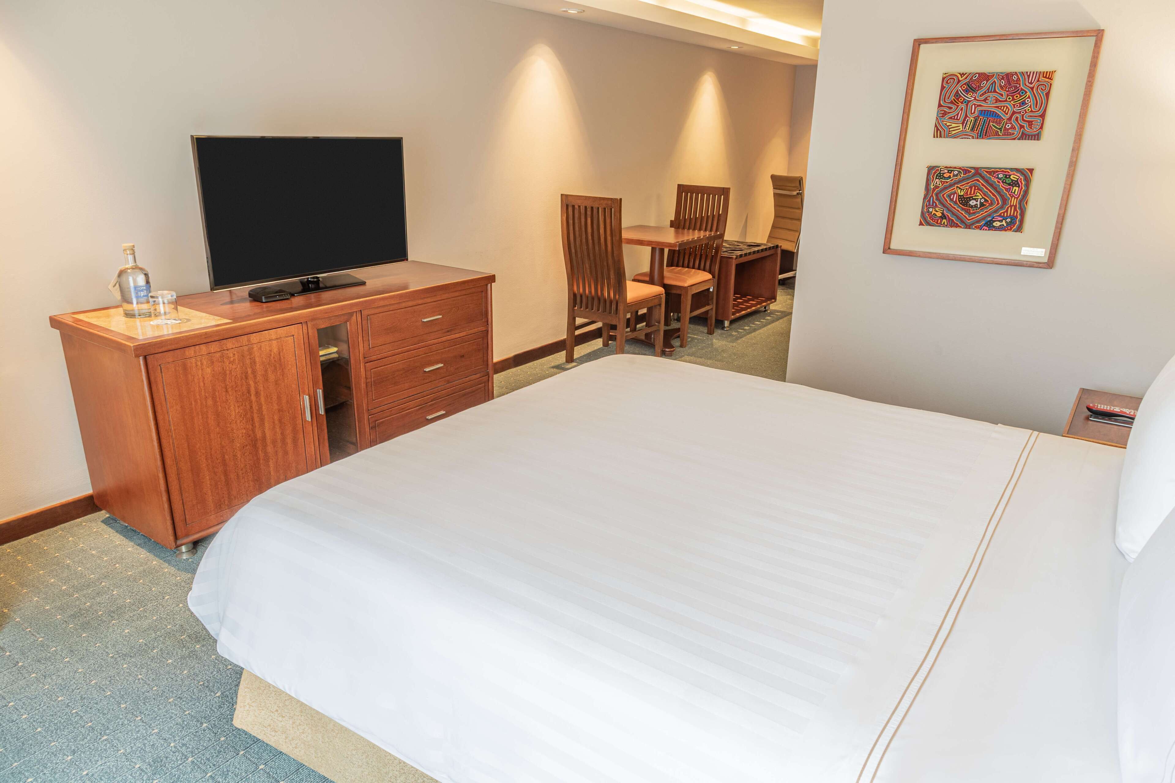 Habitación de hotel con una cama grande, un aparador de madera, un televisor de pantalla plana, un escritorio con dos sillas y cuadros en la pared.