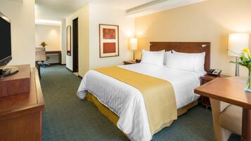 A hotel room with a large bed, a desk, a chair, a TV, and a lamp.