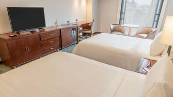 Habitación de hotel con dos camas, un escritorio, un televisor, una silla, una mesita y una ventana con vista a la ciudad.
