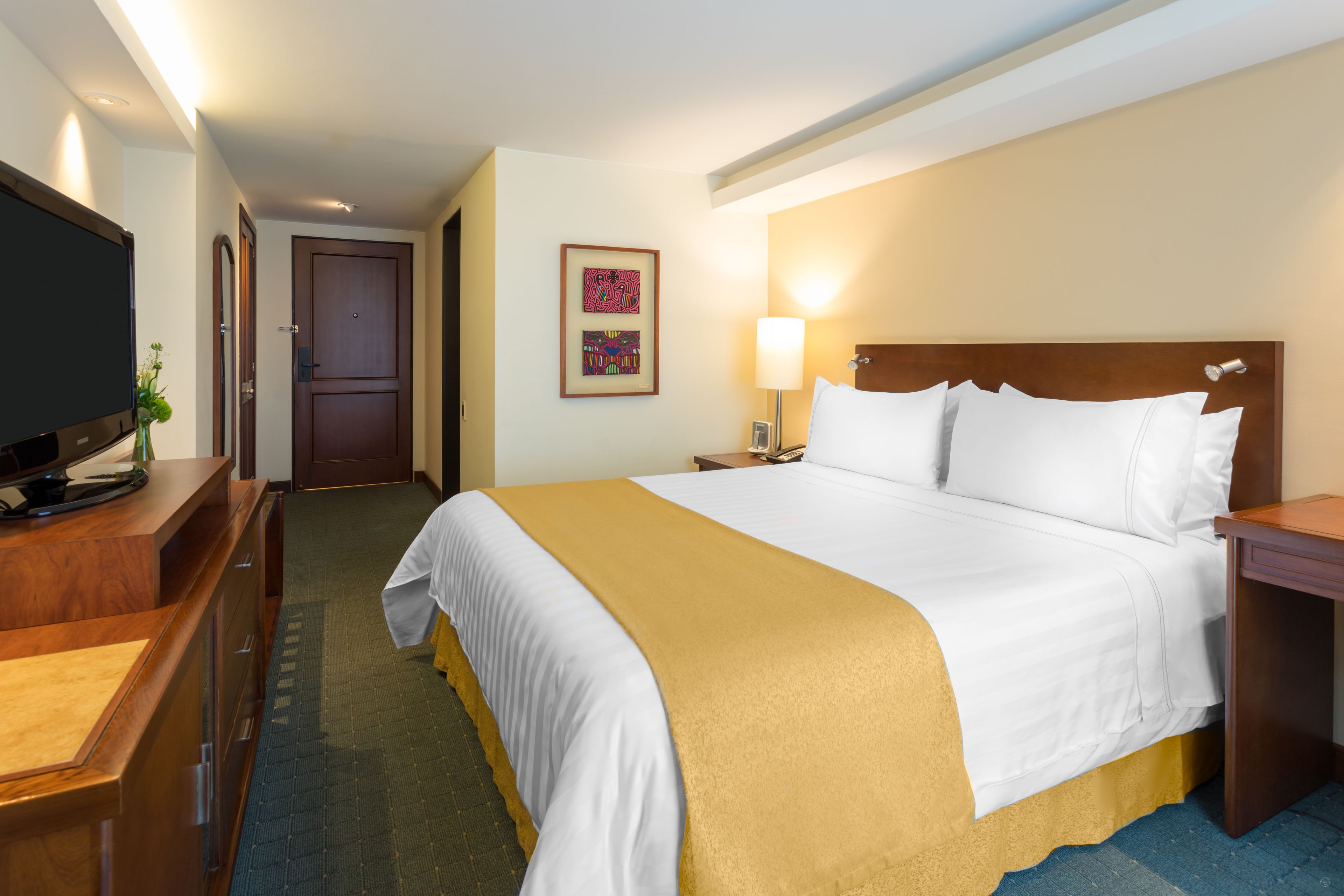 Ein Hotelzimmer mit einem großen Bett, einem Schreibtisch mit Fernseher, einer Lampe und einer Tür.