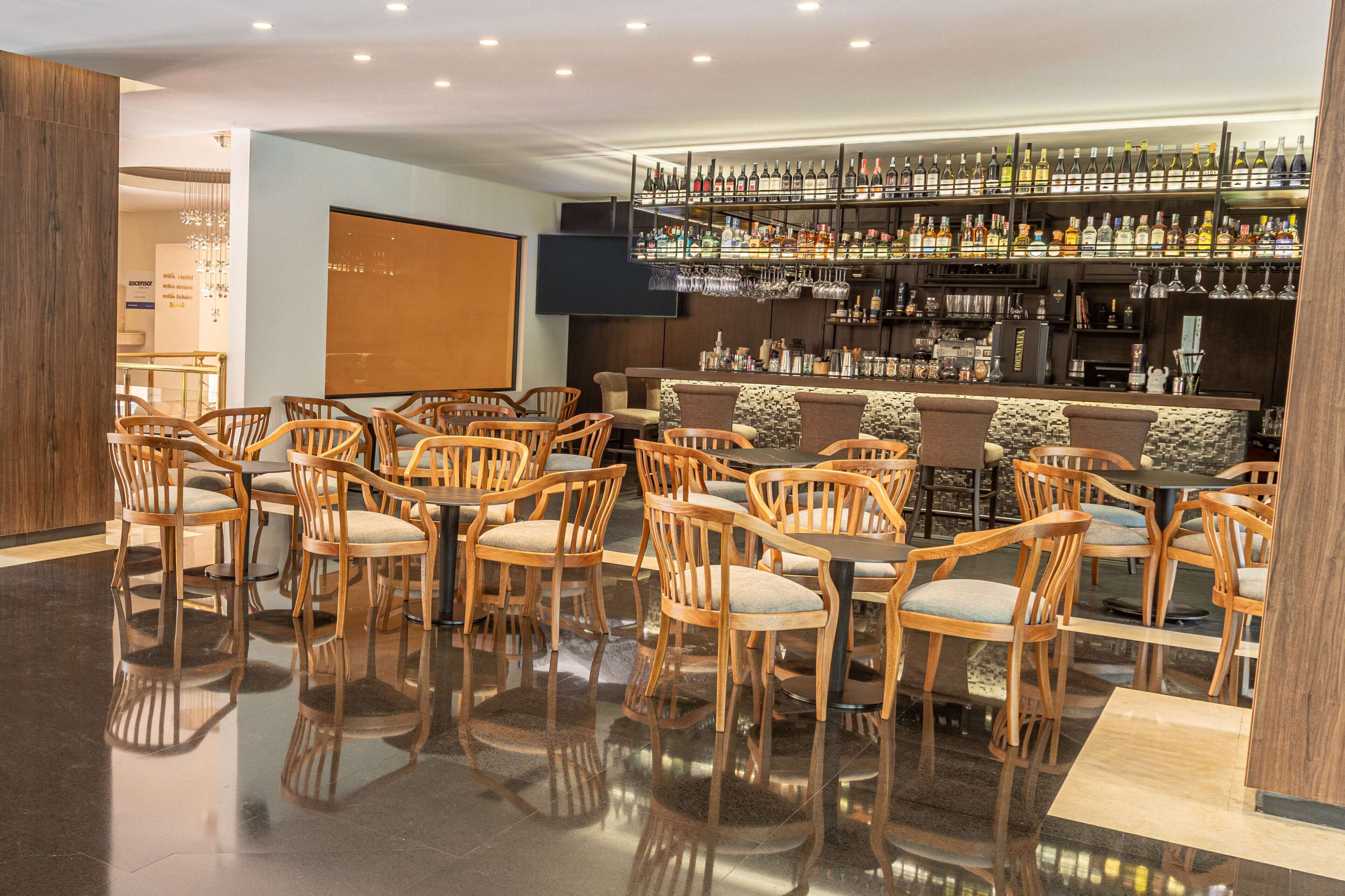 Zona de bar con sillas y mesas de madera, piso pulido y estantes con botellas de licores.