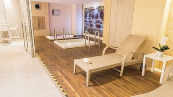 A spa room with a wooden floor, a massage table, a massage chair, a small table with a plant, and a wall-mounted clock.