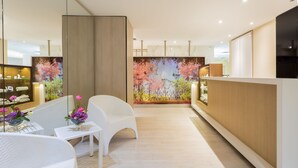 Un hall d’hôtel moderne avec un comptoir d’accueil, des fauteuils en osier blancs, une petite table agrémentée de fleurs et une grande fresque florale sur le mur.