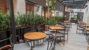 Un café en plein air avec des tables rondes en bois et des chaises en métal, entouré de jardinières et d’un mur de briques.