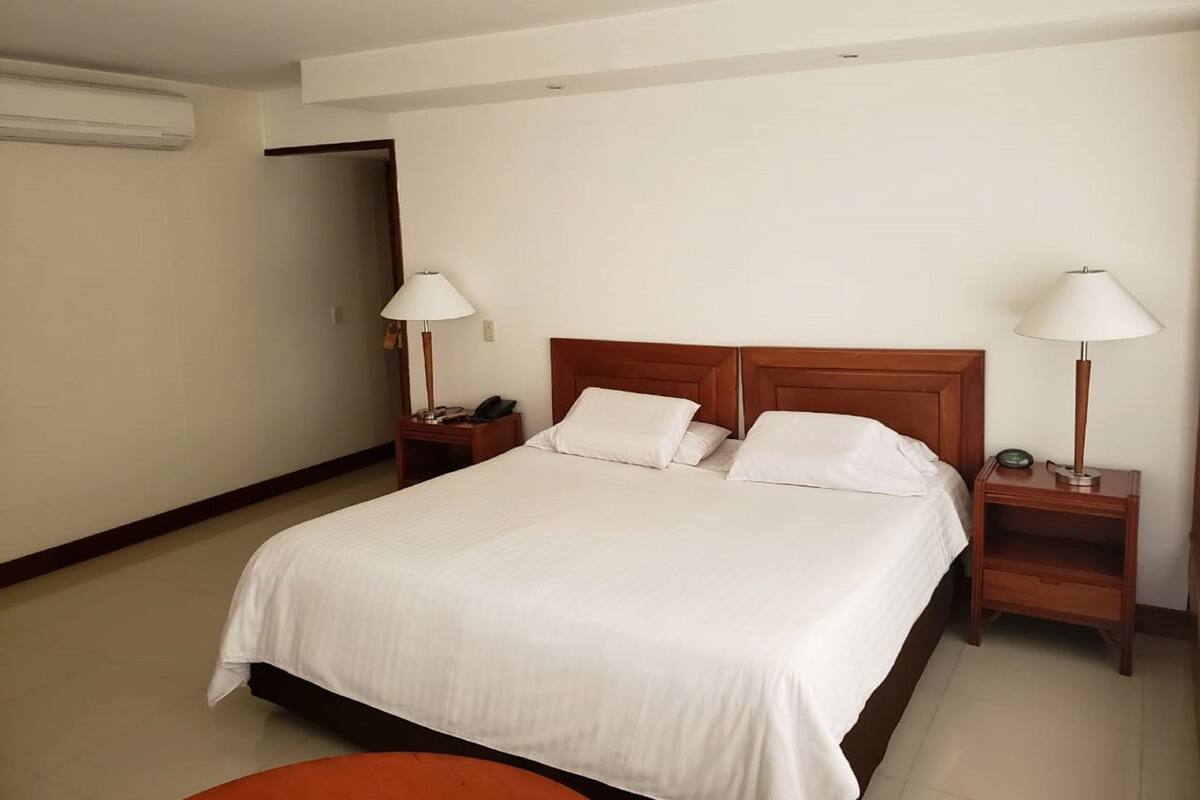Une chambre d’hôtel avec un grand lit, deux tables de chevet, une petite table, une lampe et un climatiseur fixé au mur.