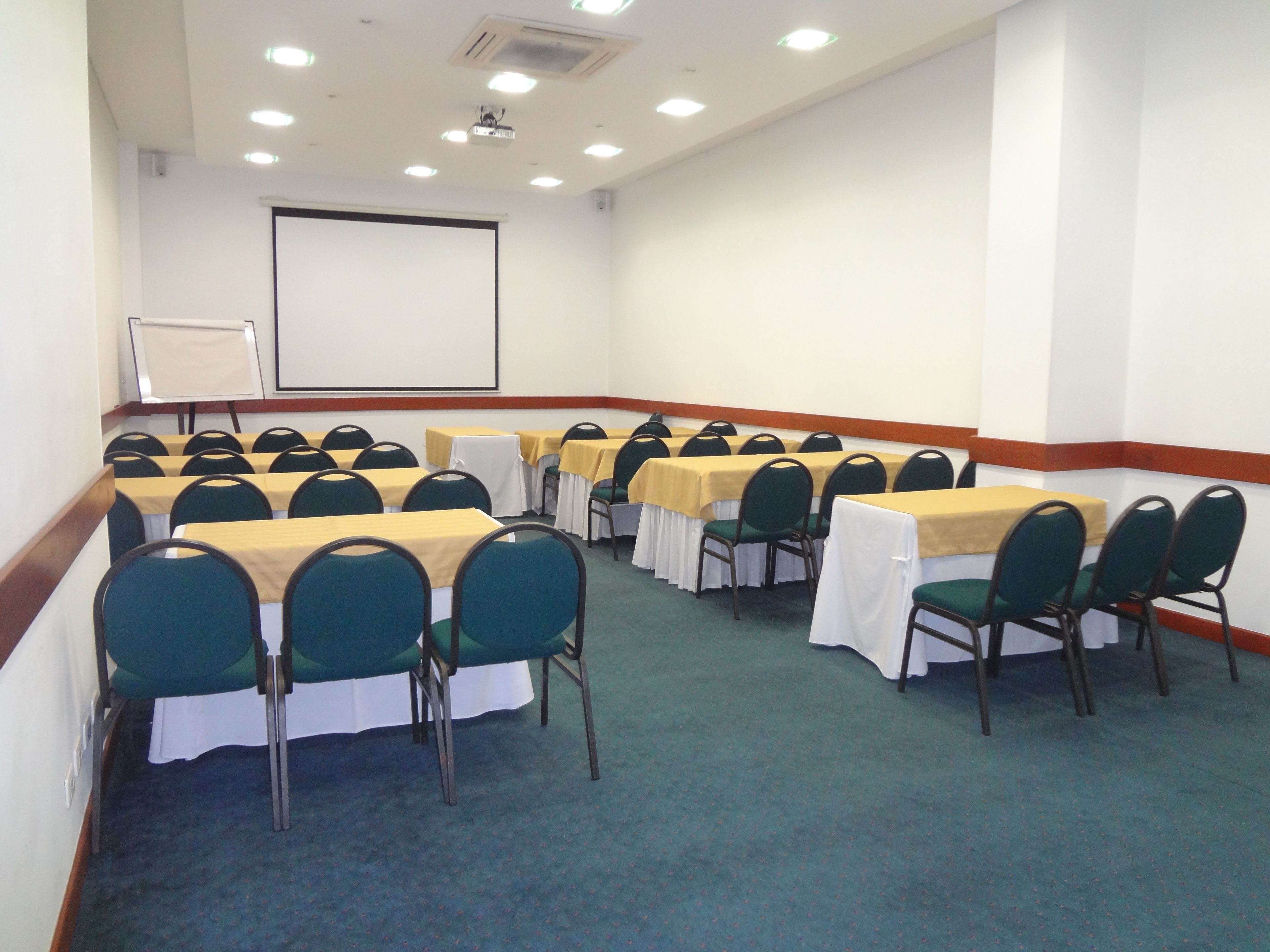 Uma sala de conferência com fileiras de mesas e cadeiras, uma tela de projetor e um quadro branco.
