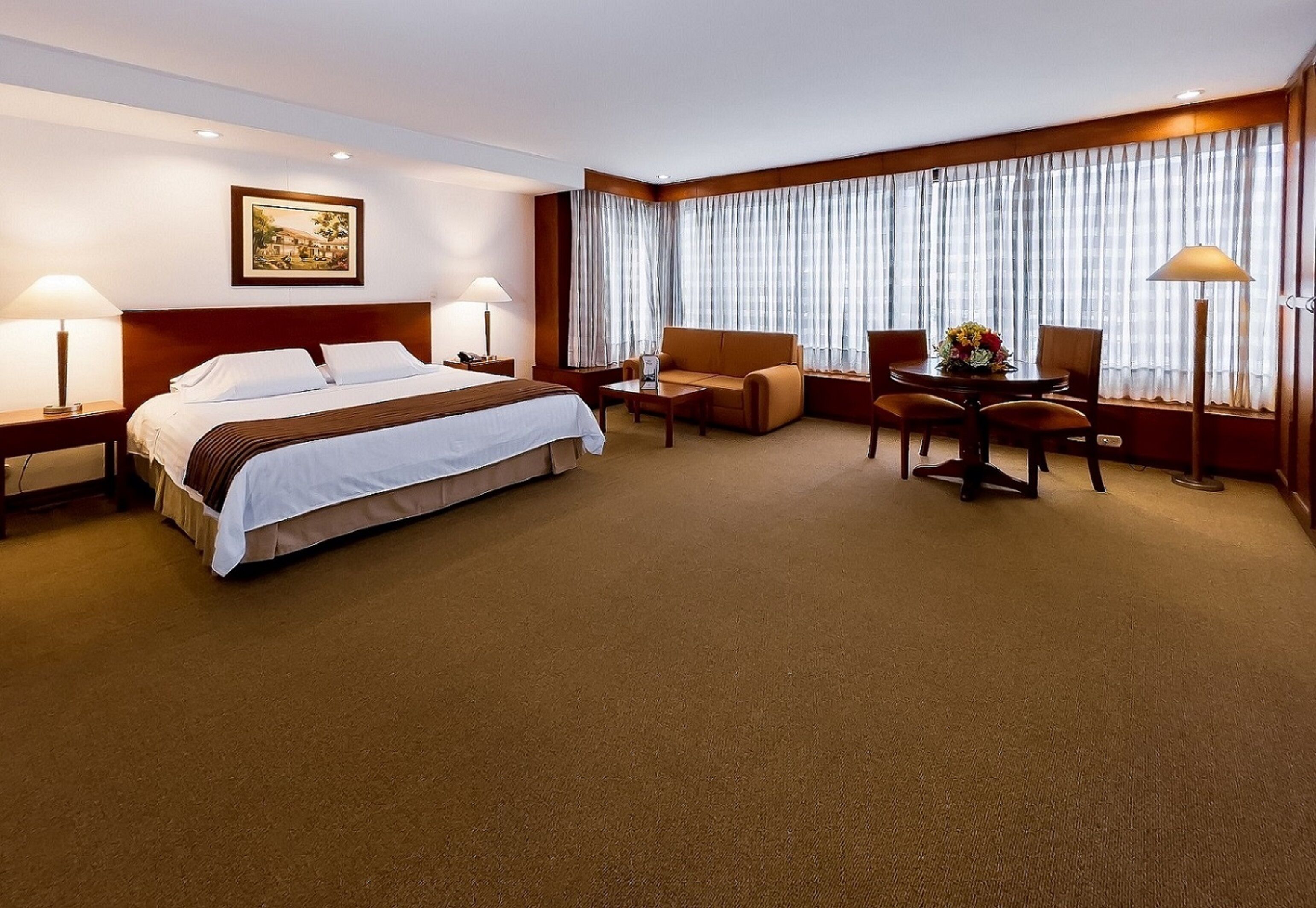 A hotel room with a large bed, a sofa, a dining table with chairs, and a large window with curtains.