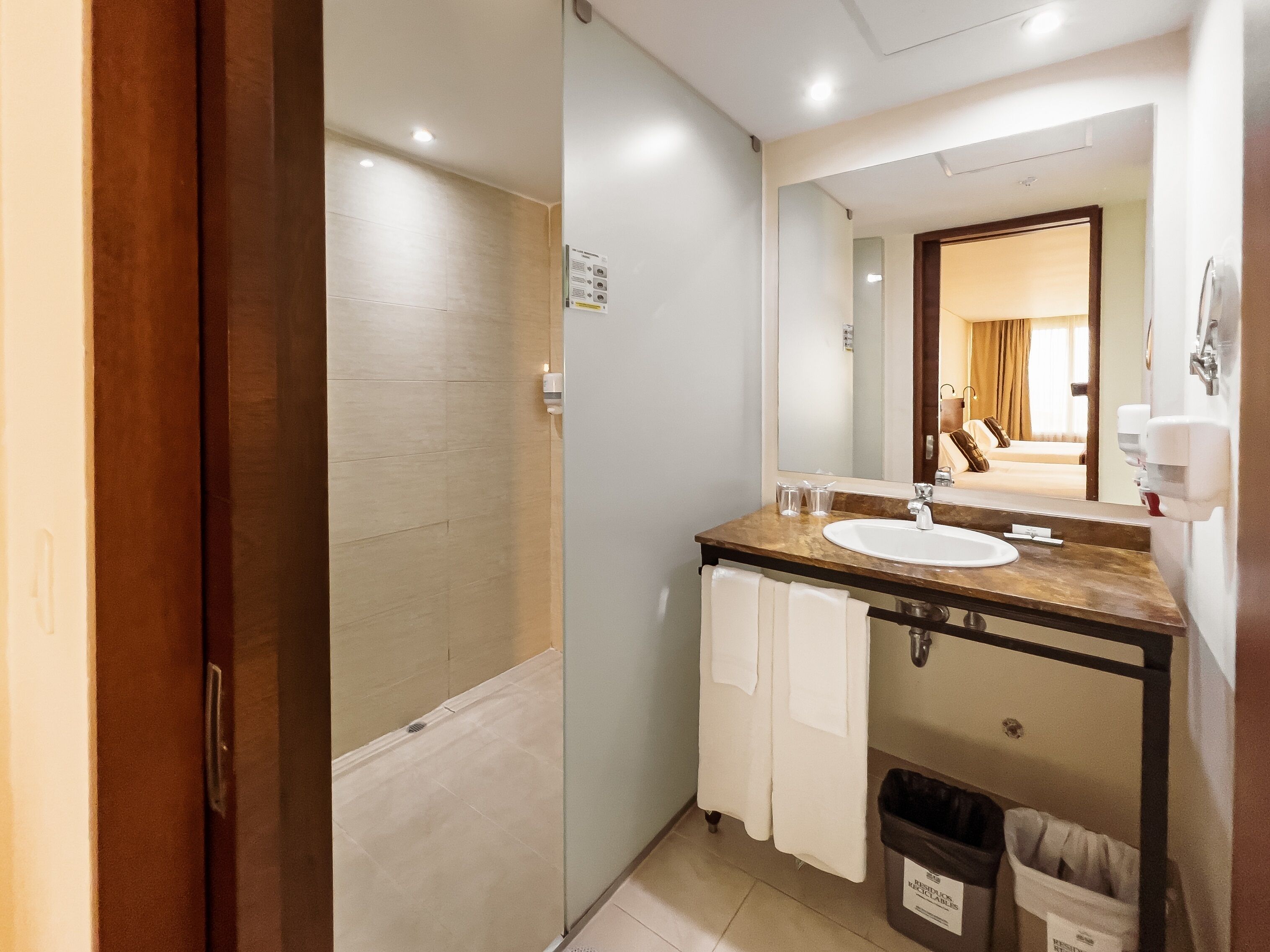 A bathroom with a shower, sink, and mirror. There is a trash bin and a towel rack.