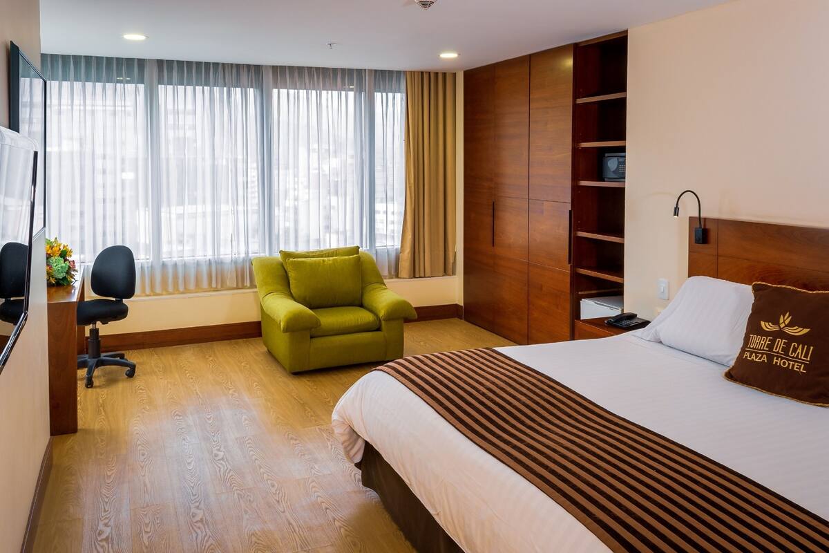Une chambre d’hôtel avec un lit, un fauteuil vert, un bureau avec un ordinateur et une grande fenêtre avec des rideaux.