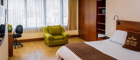 Una habitación de hotel con una cama, un sillón verde, un escritorio con una computadora y un ventanal con cortinas.