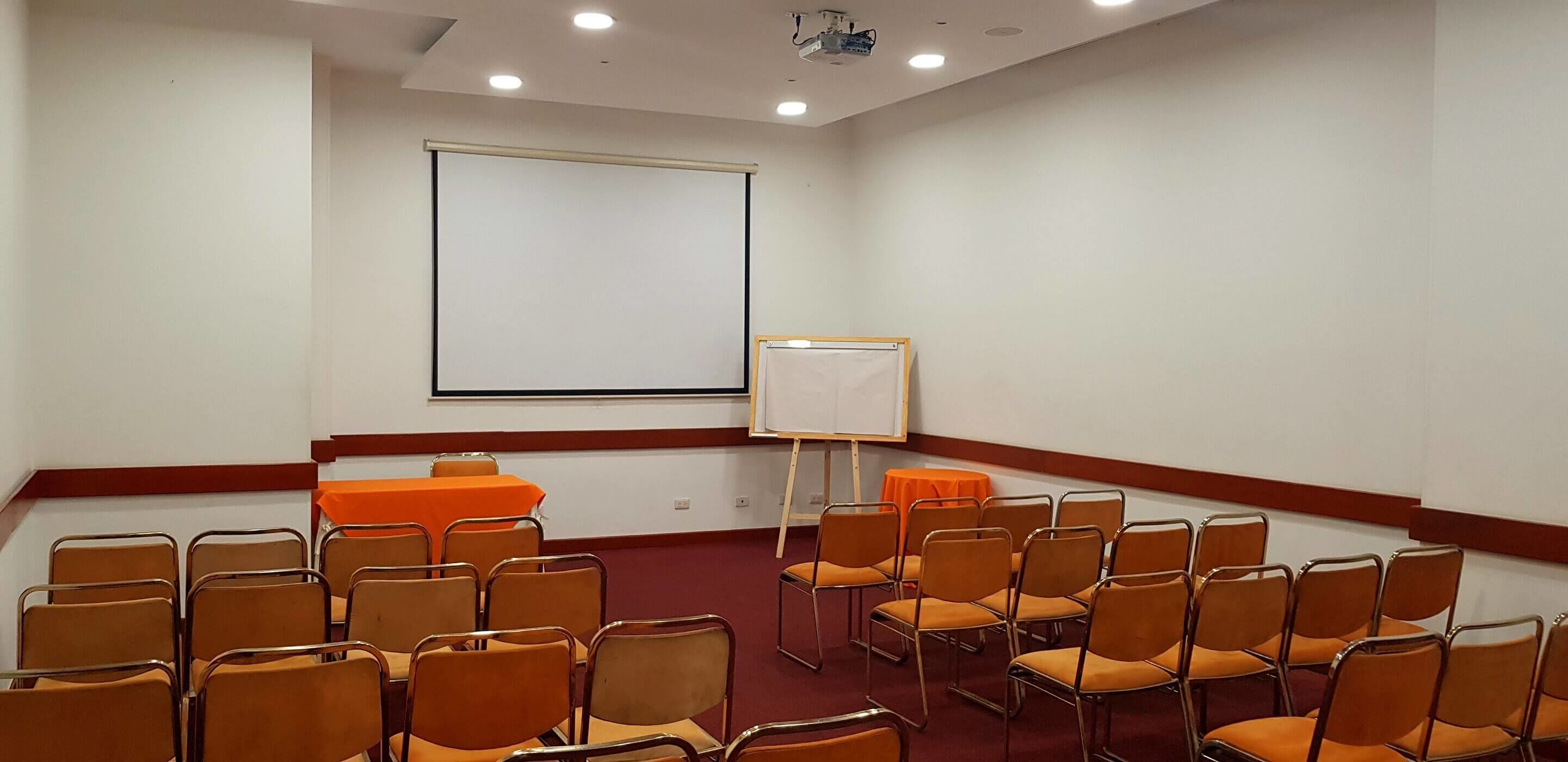 Un salón de conferencias con una pantalla grande, una pizarra y filas de sillas naranjas.