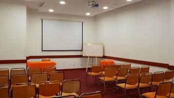 Un salón de conferencias con una pantalla grande, una pizarra y filas de sillas naranjas.