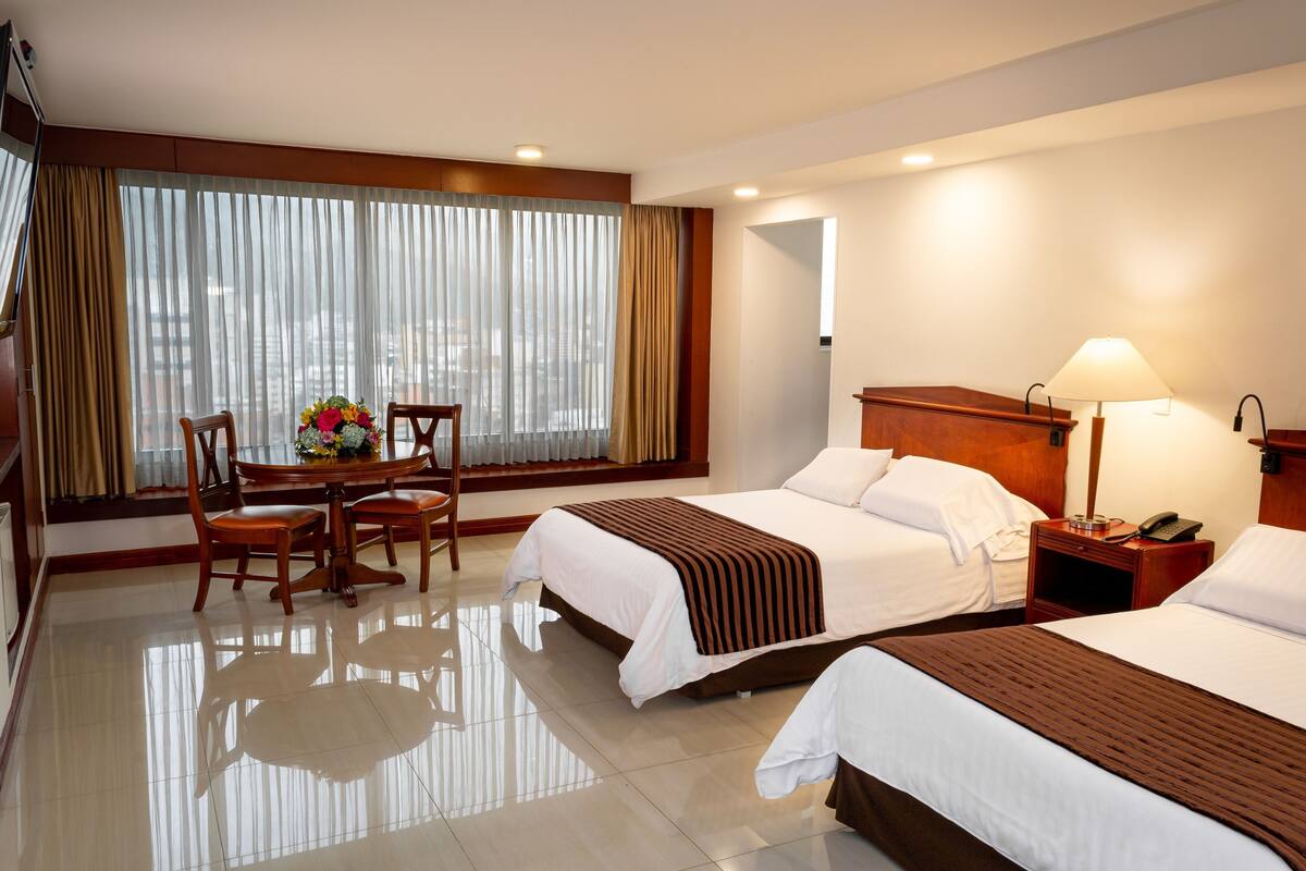 Une chambre d’hôtel avec deux lits, une table à manger avec des chaises, une grande fenêtre avec des rideaux et une table de chevet avec une lampe et un téléphone.