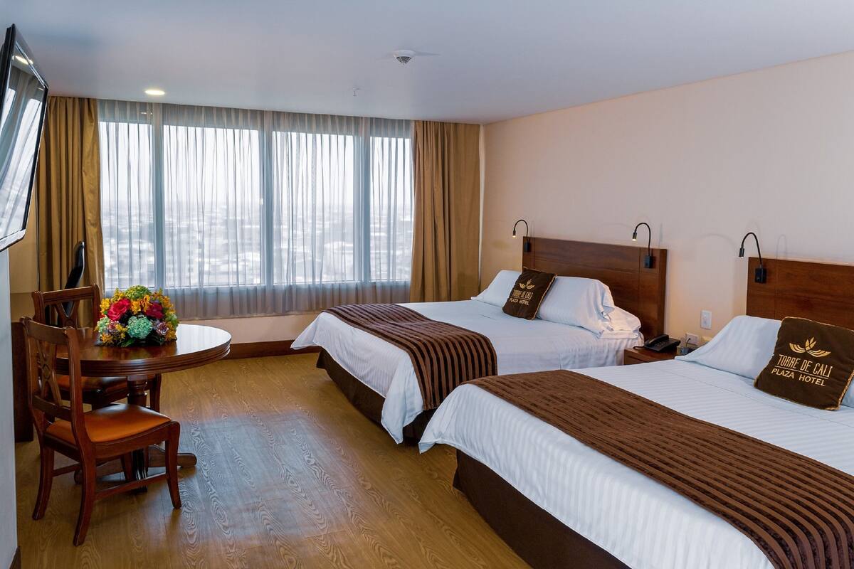 Une chambre d’hôtel avec deux lits, une table à manger avec des chaises et une grande fenêtre avec des rideaux.