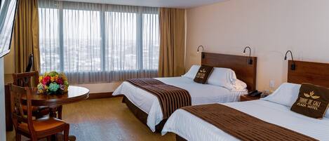 Ein Hotelzimmer mit zwei Betten, einem Esstisch mit Stühlen und einem großen Fenster mit Vorhängen.
