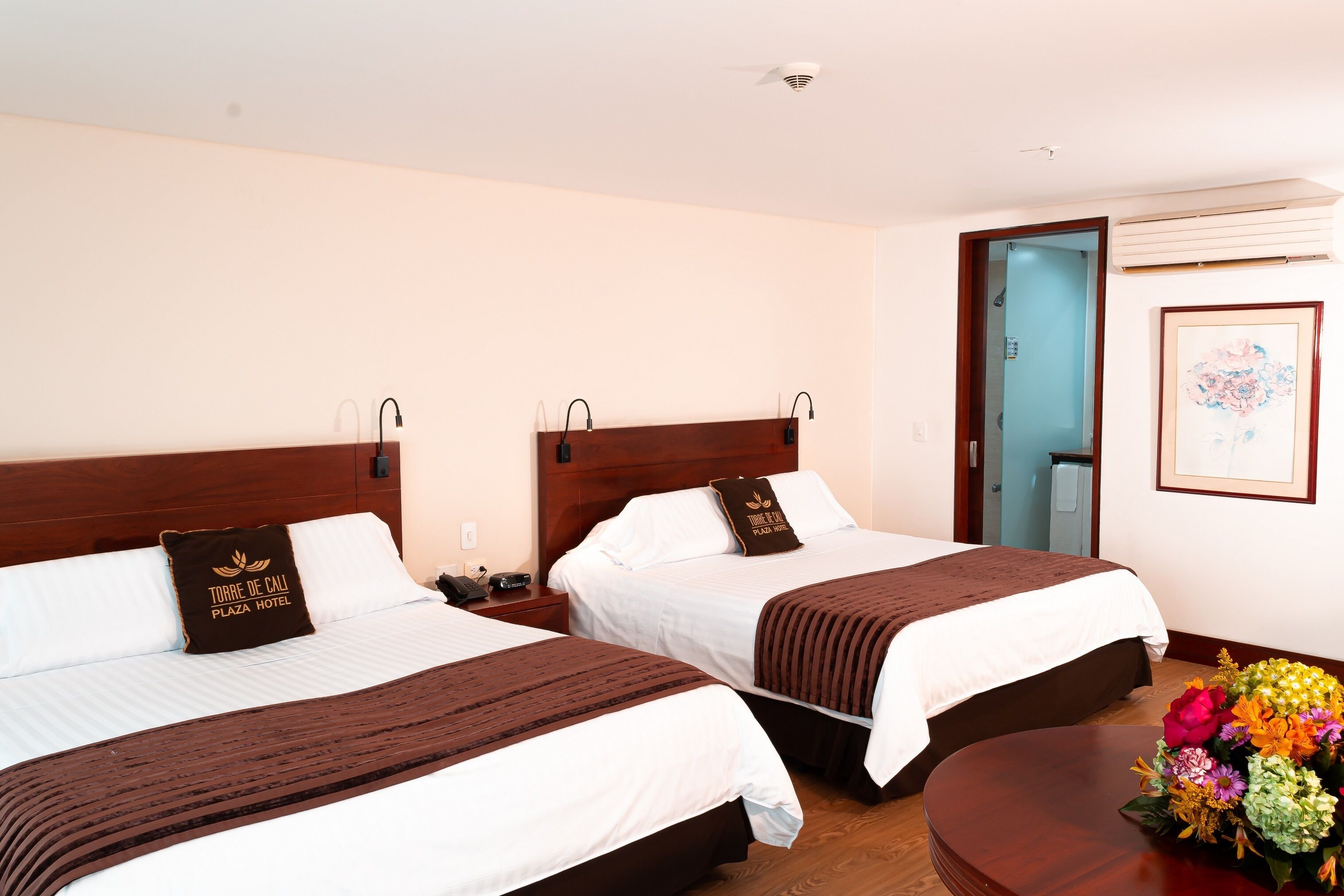 Quarto de hotel com duas camas, cada uma com cabeceira marrom e almofada com o nome do hotel. Há uma mesa de cabeceira com telefone e abajur, e uma mesa de madeira com um buquê de flores.