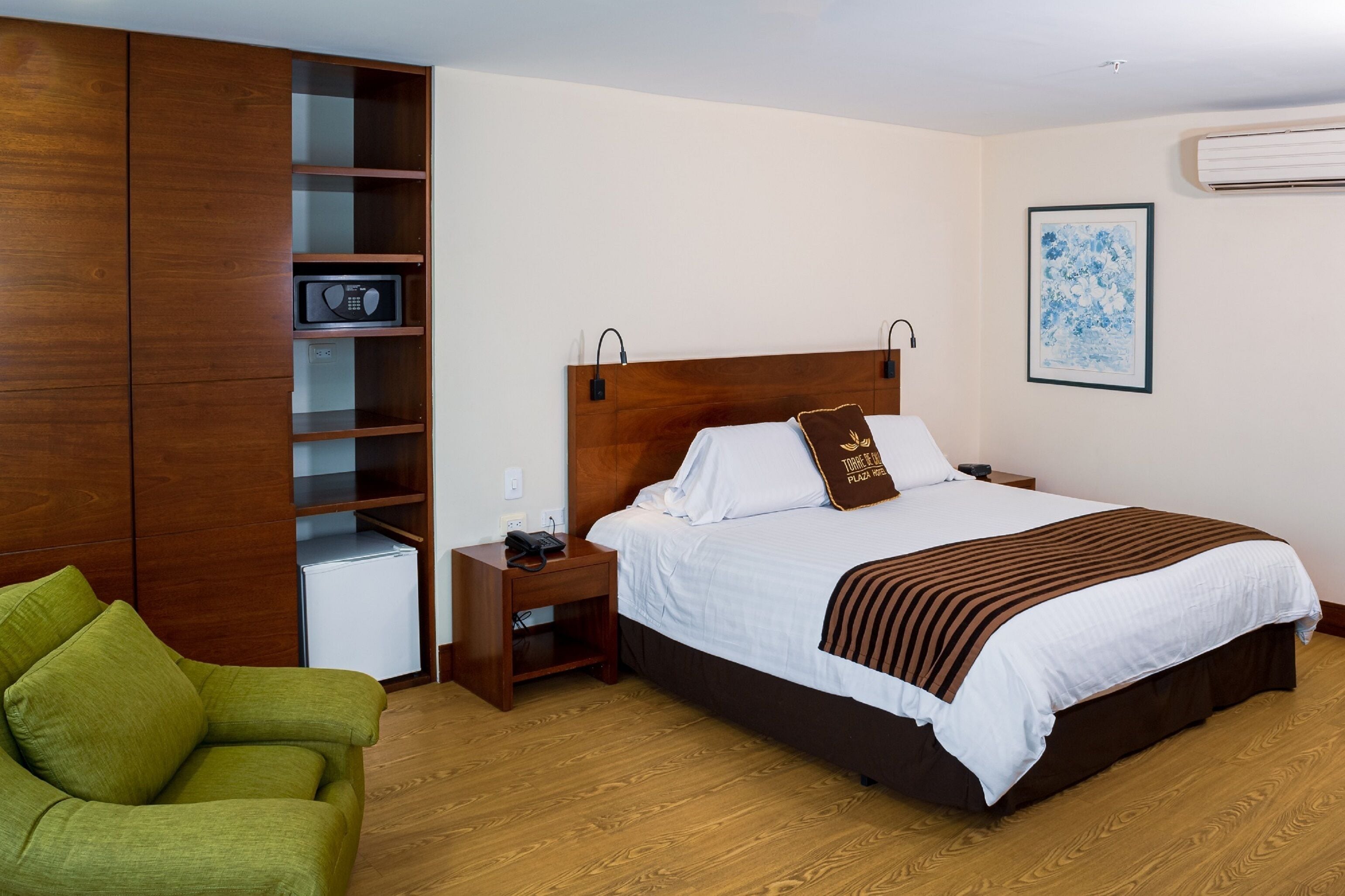 A hotel room with a large bed, a wooden wardrobe, a small desk with a phone, a green armchair, and a framed artwork on the wall.