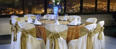 A banquet table set for a formal event with chairs adorned with gold sashes, a floral centerpiece, and a cityscape view through large windows.