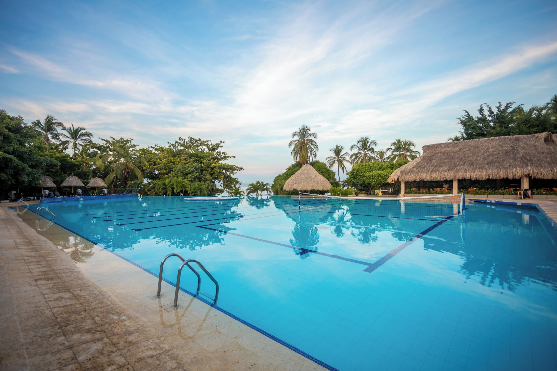A large swimming pool with clear blue water, surrounded by palm trees and thatched-roof huts.