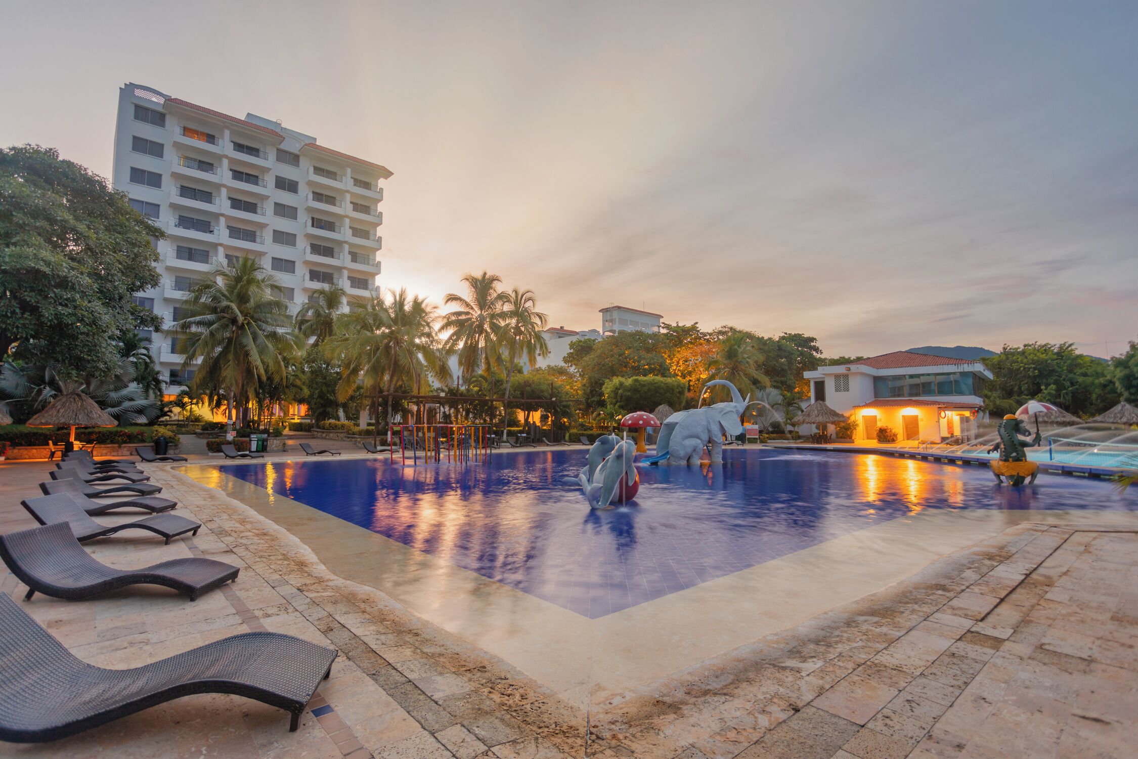 Una piscina con sillas de descanso, palmeras y un edificio de hotel al fondo.