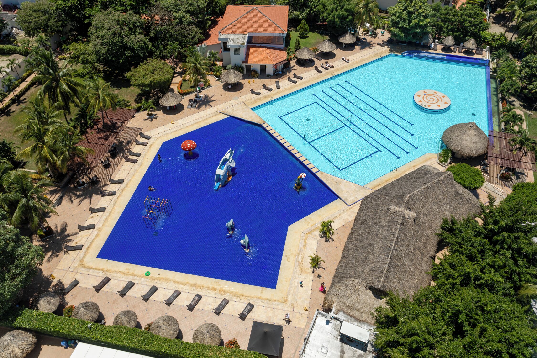 An aerial view of a resort with two swimming pools, a beach area, and a building with a red roof.