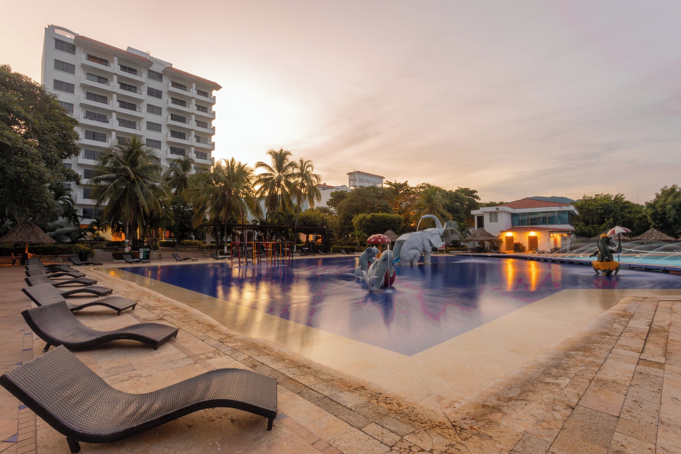 A swimming pool area with lounge chairs, palm trees, and a building in the background.