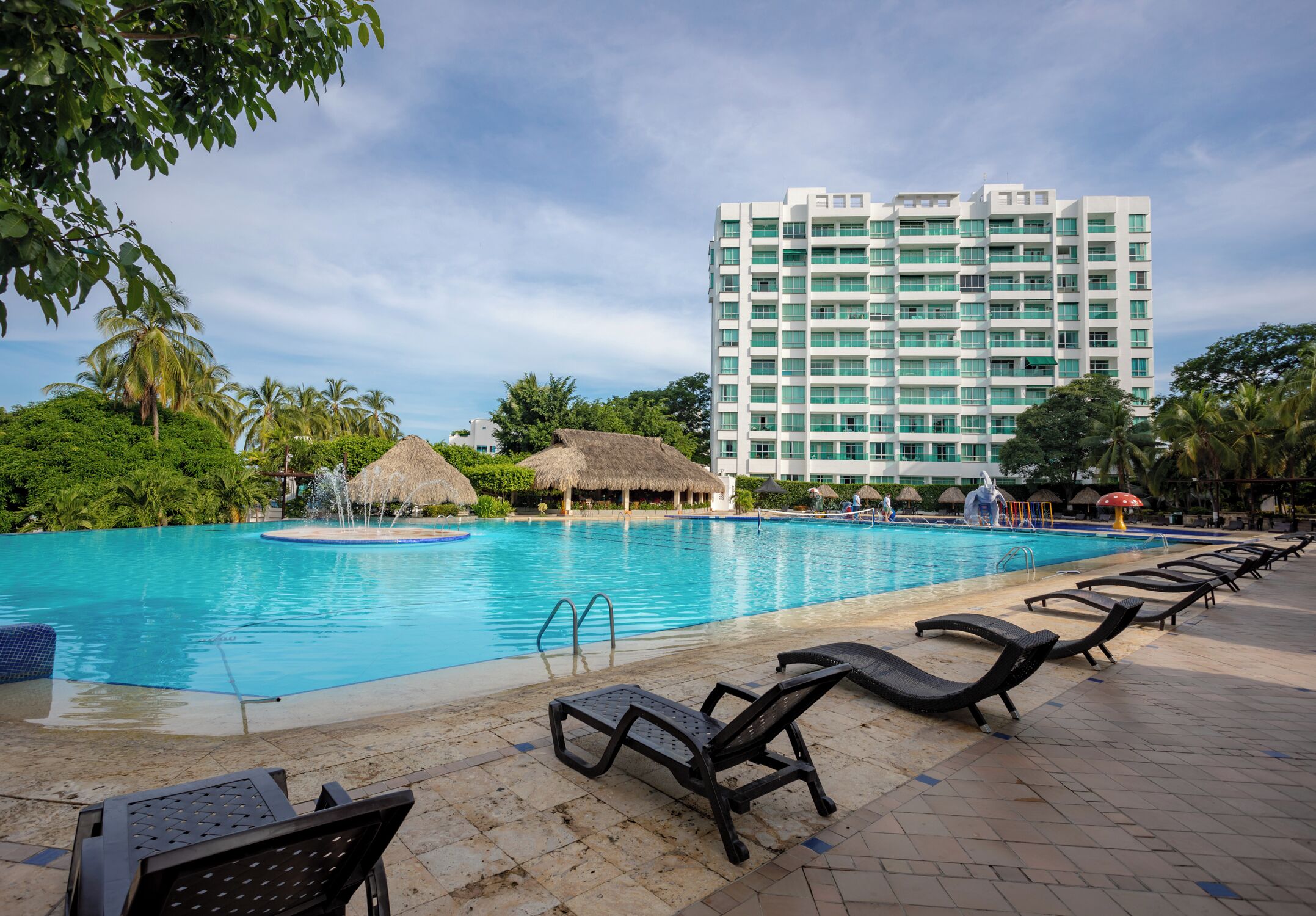 A swimming pool with lounge chairs, a thatched-roof structure, and a tall hotel building in the background.