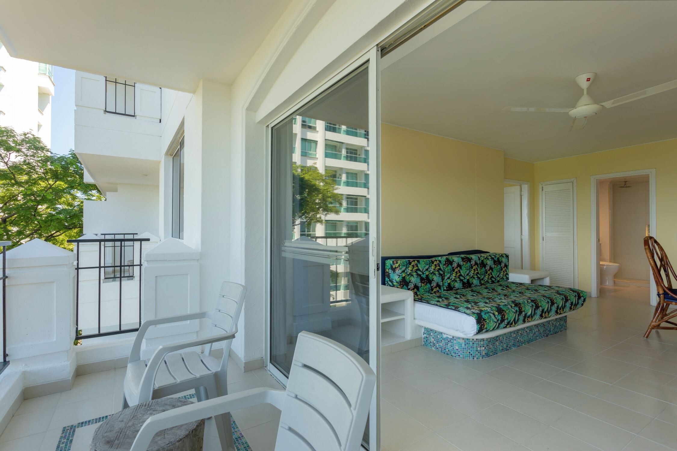 A balcony with white chairs and a table, a sliding glass door leading to a room with a bed and a chair, and a ceiling fan.
