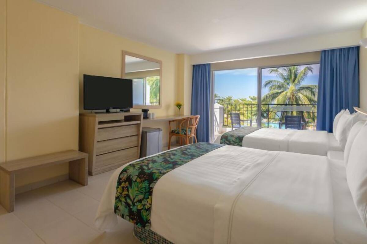 Habitación de hotel con dos camas, televisión, escritorio y balcón con vista a palmeras.