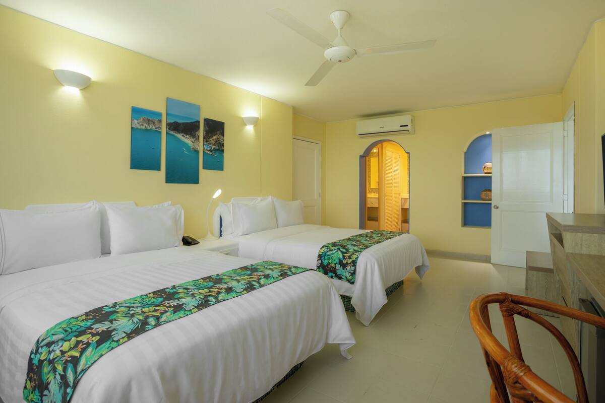 Habitación de hotel con dos camas, un escritorio, una silla, ventilador de techo, obras de arte en la pared y un equipo de aire acondicionado.
