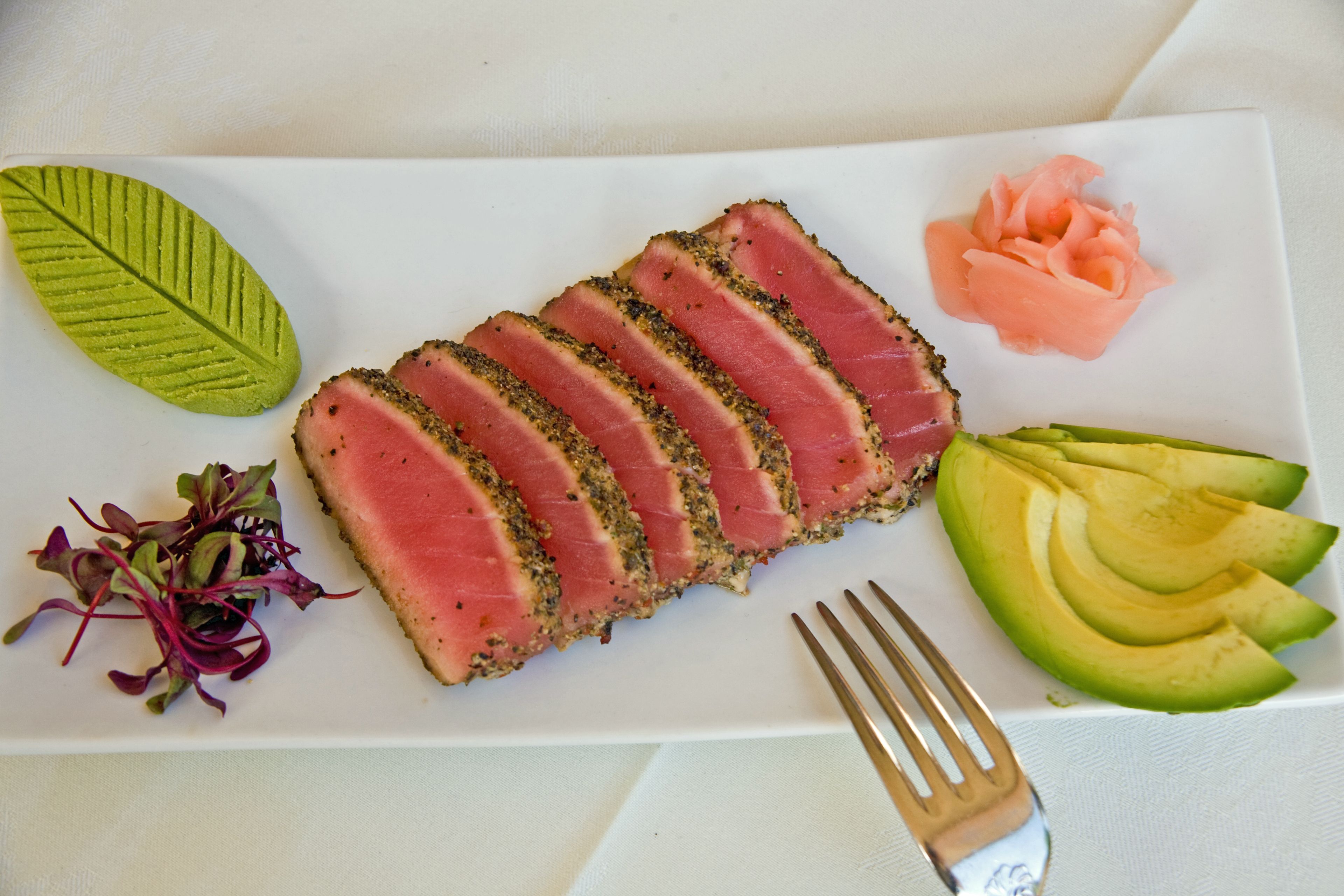 A white rectangular plate with sliced tuna, avocado, and a green leaf garnish.