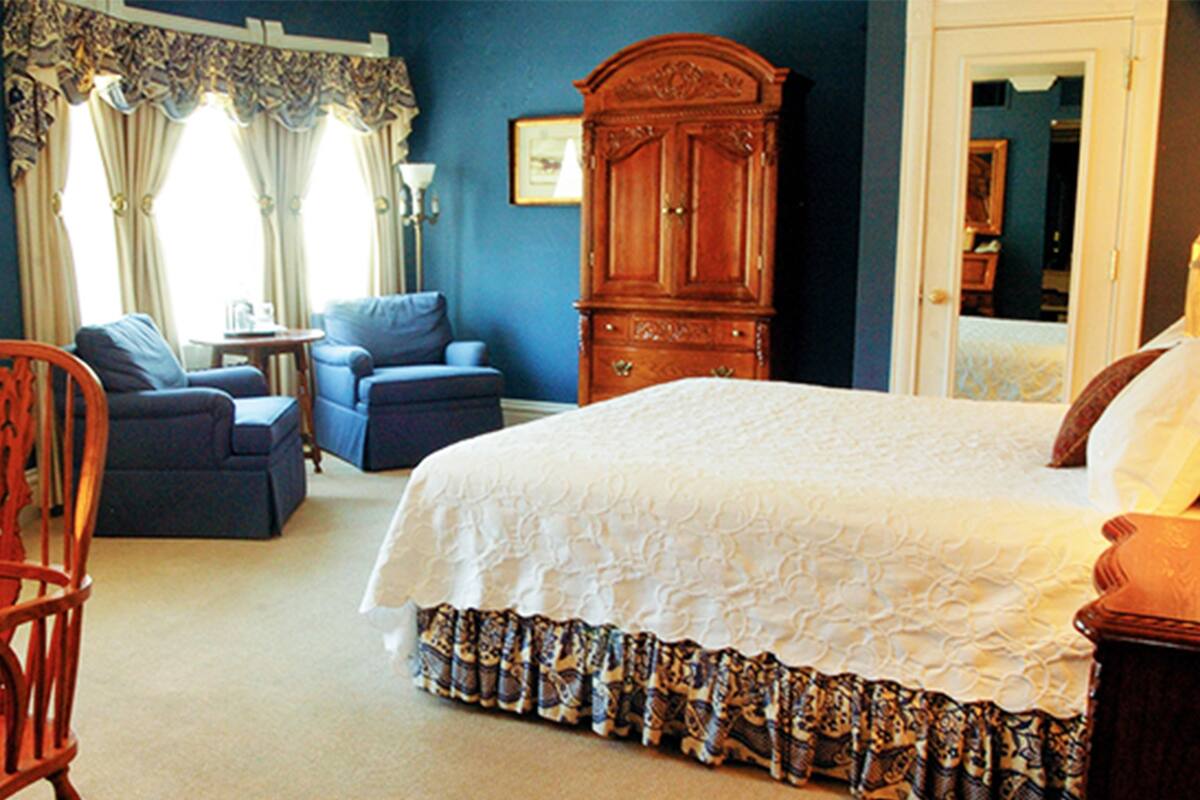 A bedroom with a large bed, two armchairs, a wooden cabinet, and a door leading to another room.