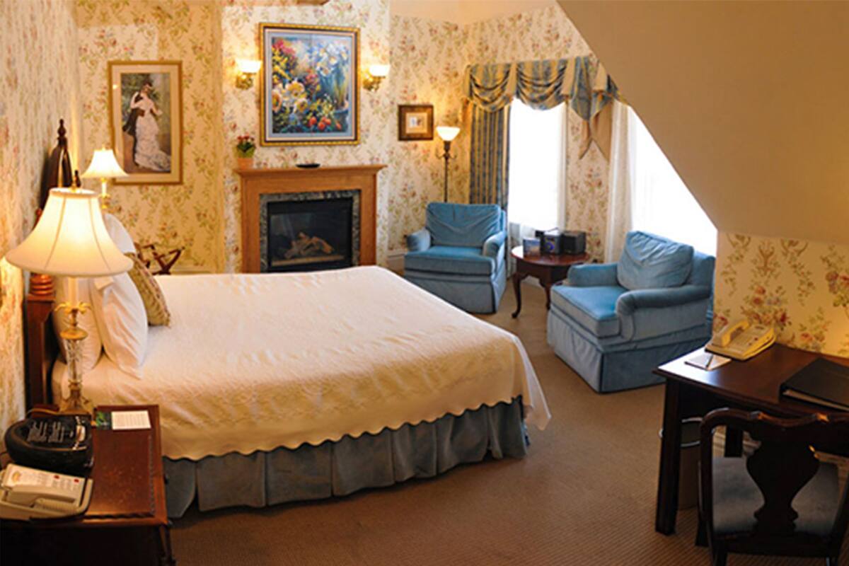 A hotel room with a large bed, a chair, a small table, a lamp, a fireplace, and a painting on the wall.