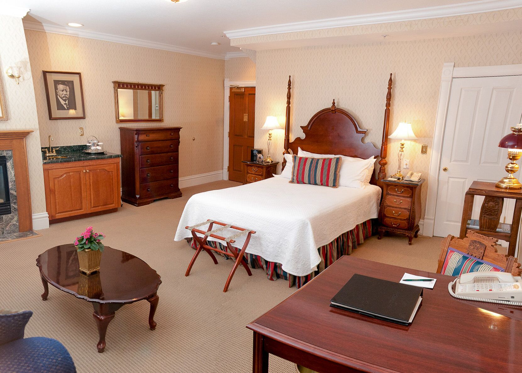 Une chambre d’hôtel avec un grand lit, une commode en bois, un bureau avec un téléphone, une chaise et une petite table sur laquelle est posé un vase.