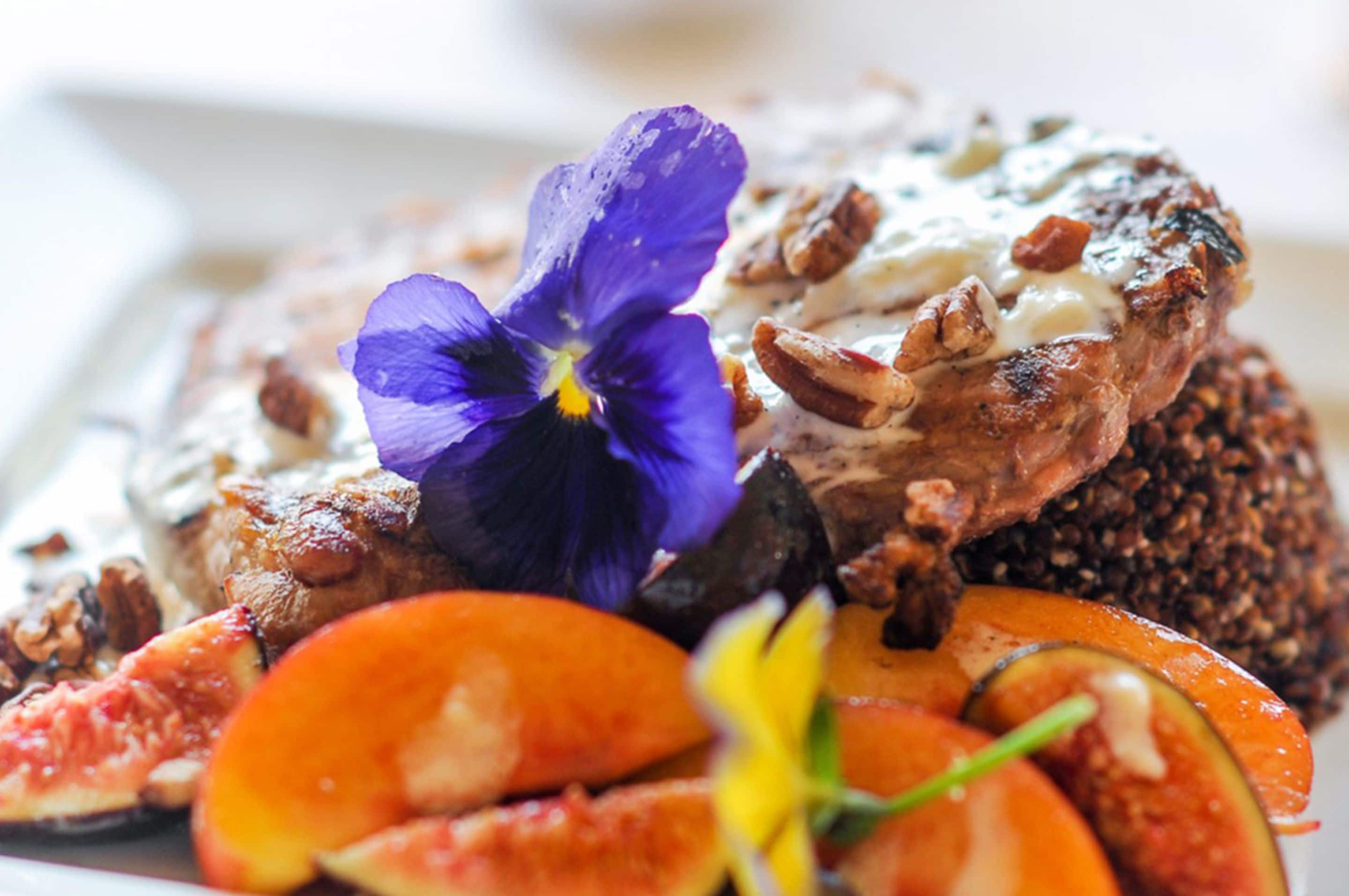Une assiette contenant un morceau de viande nappé de sauce et parsemé de noix, accompagné de légumes coupés en tranches et d’une fleur.