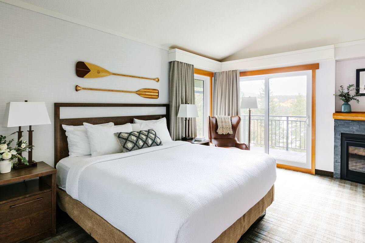 Habitación de hotel con una cama grande, una mesita de noche con lámpara, una silla, chimenea y balcón con vistas.