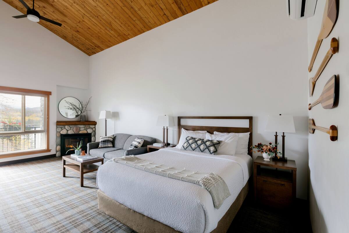 Un dormitorio con una cama grande, techo de madera, chimenea, sofá y mesita de noche con lámpara.