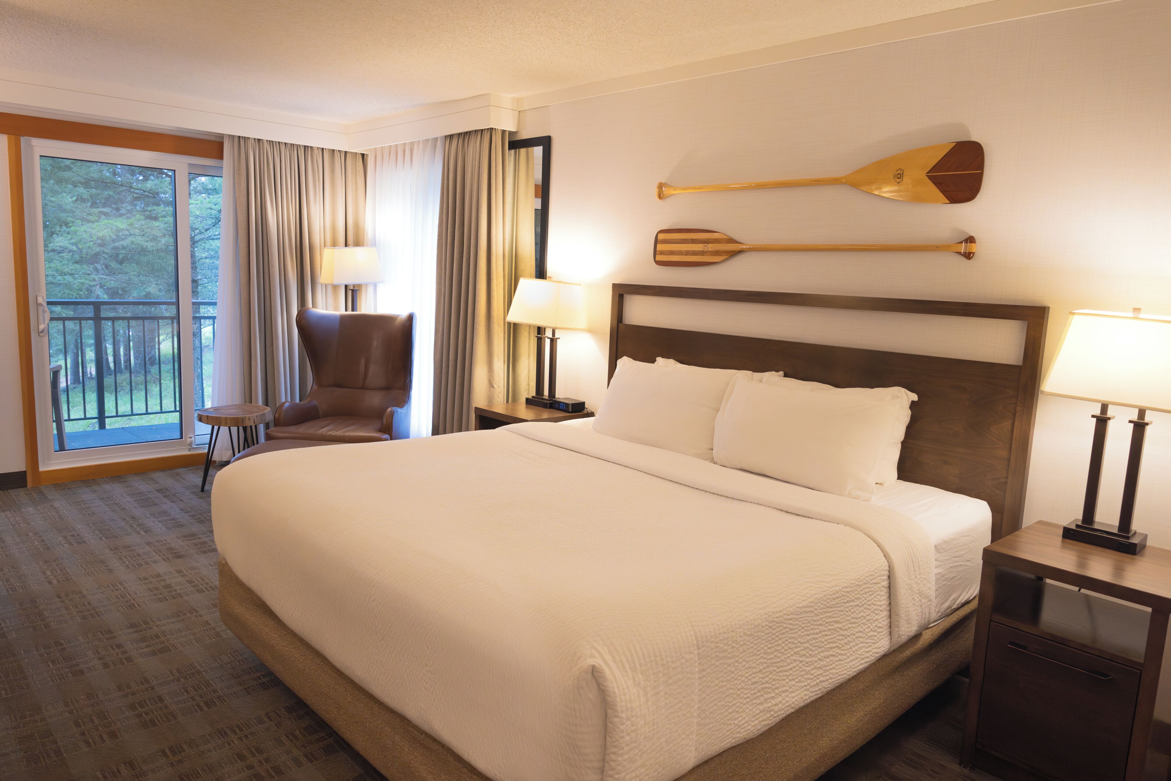 A hotel room with a large bed, a chair, a nightstand, a lamp, a window with curtains, and a wall-mounted paddle.