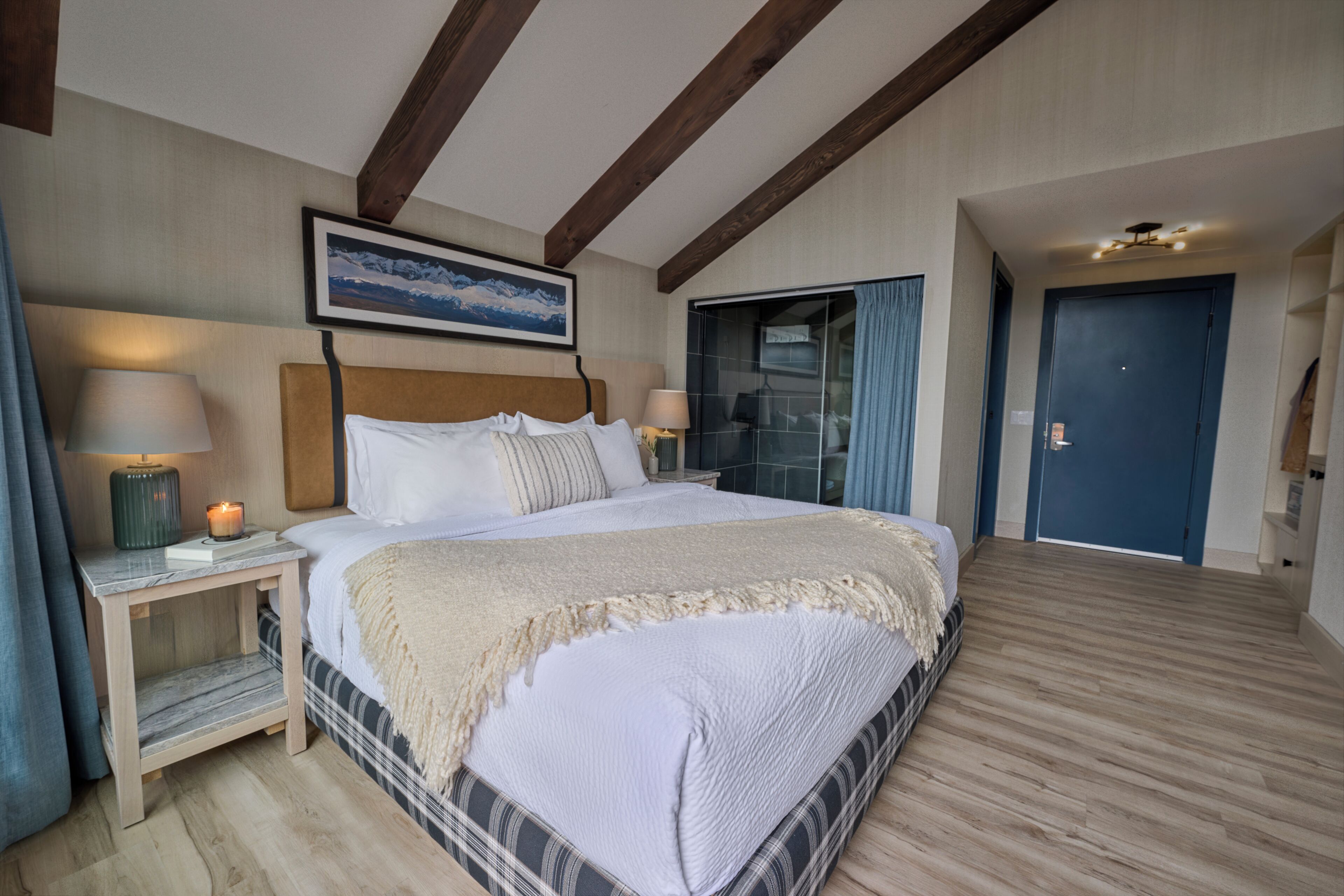 Un dormitorio con cama, mesitas de noche, lámpara, un cuadro, una puerta y una ventana.
