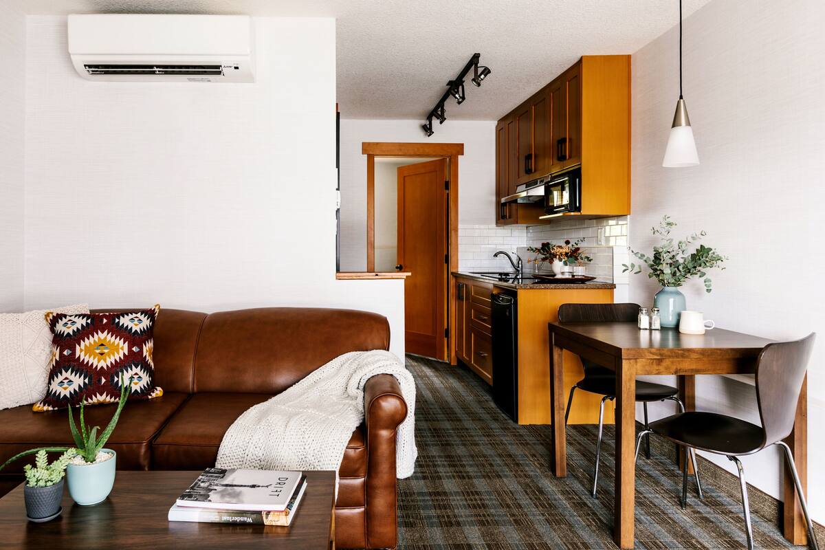 Una sala moderna con un sofá de cuero marrón, una mesa de centro con una revista y una planta en maceta. En el fondo, se ve una mesa de comedor de madera con sillas.