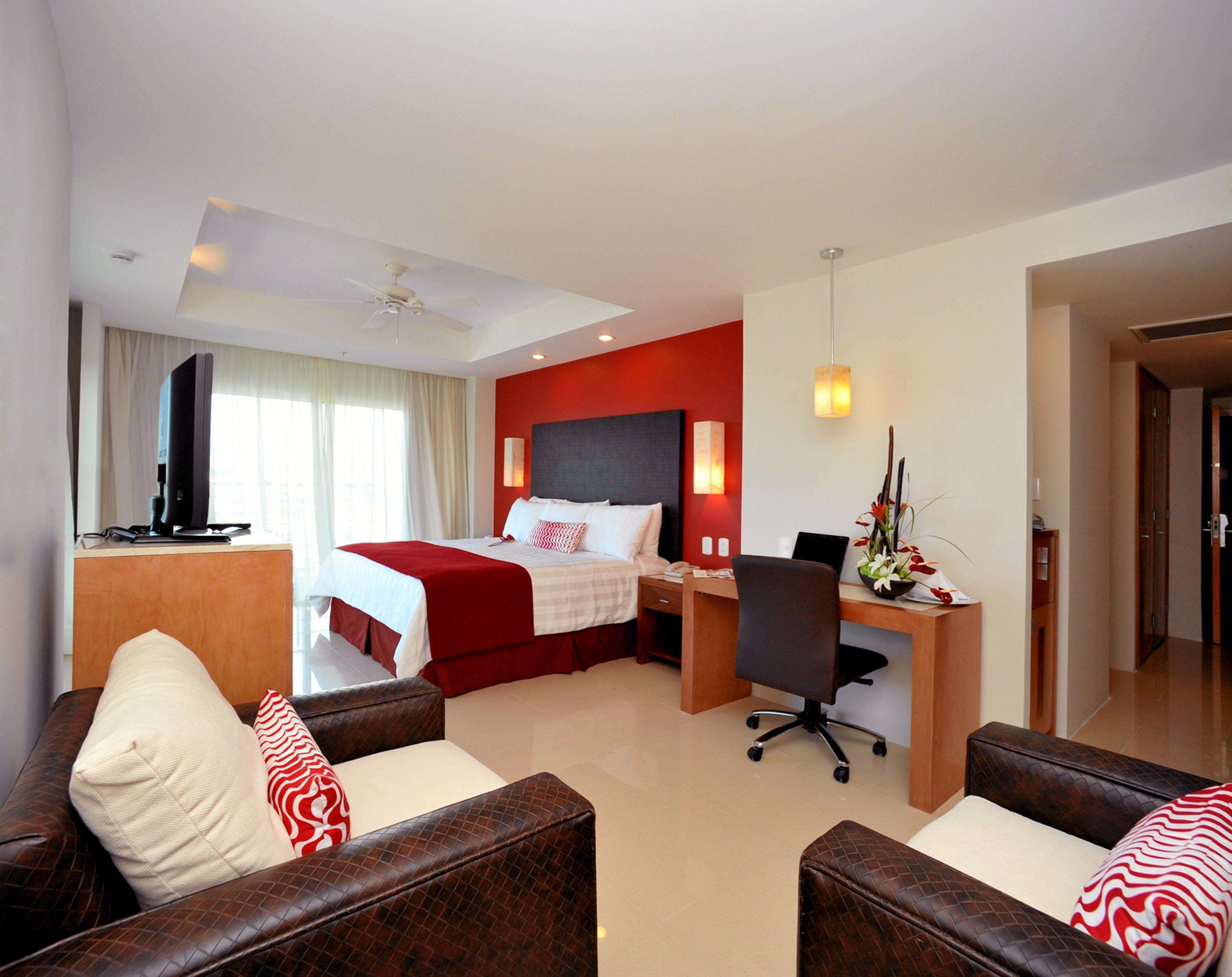 A hotel room with a bed, a desk, two leather sofas, a television, and a large window with curtains.