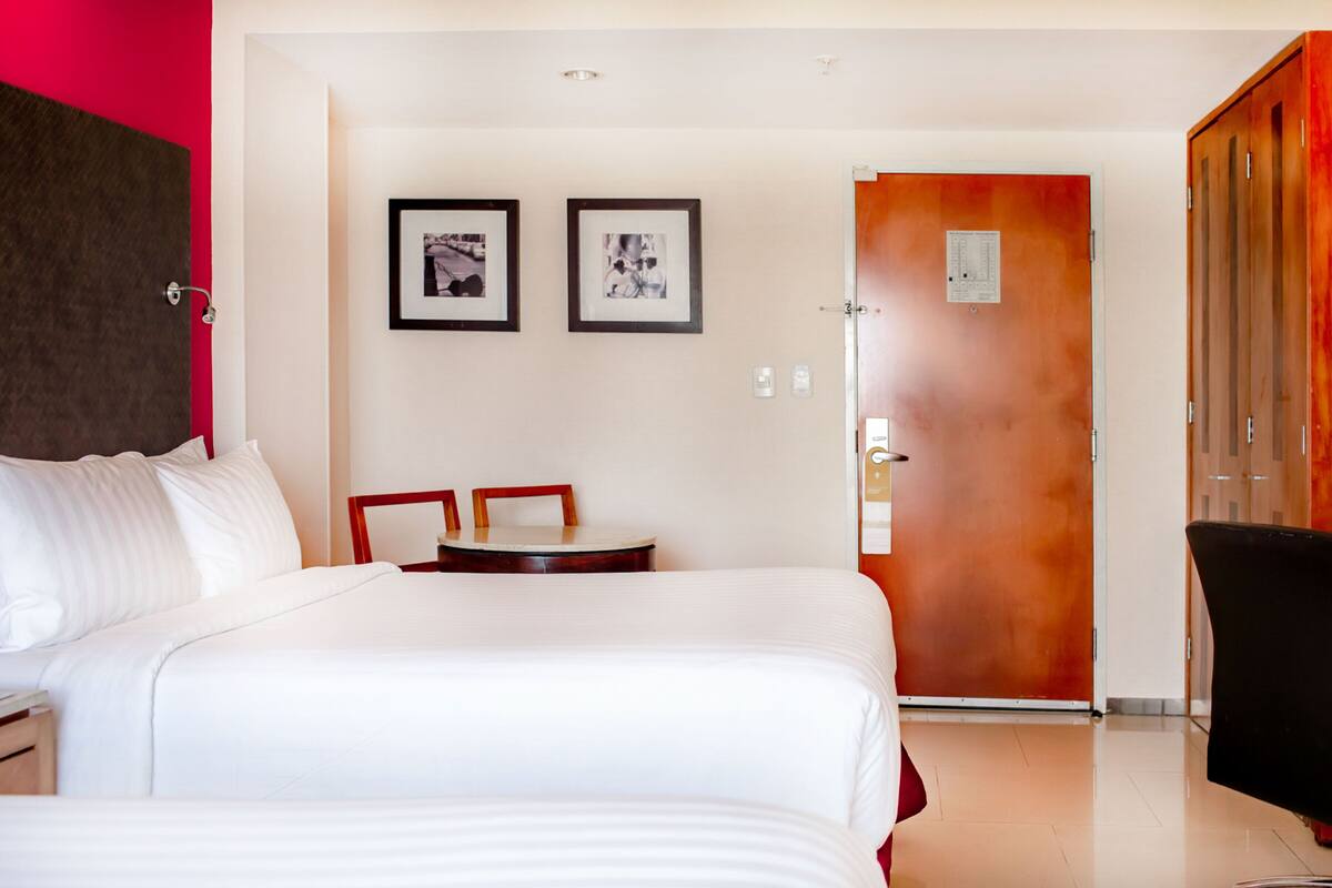 Habitación de hotel con una cama grande, una mesita pequeña, dos cuadros en la pared y una puerta que da a otra habitación.