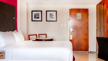 A hotel room with a large bed, a small table, two framed pictures on the wall, and a door leading to another room.