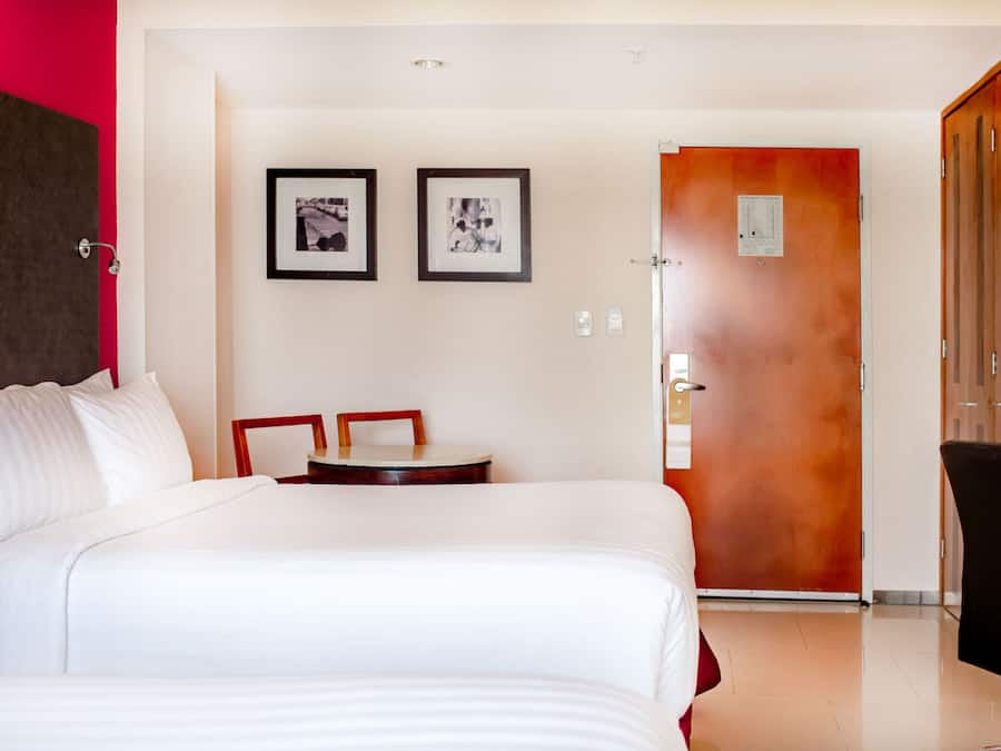 A hotel room with a large bed, a small table, two framed pictures on the wall, and a door leading to another room.
